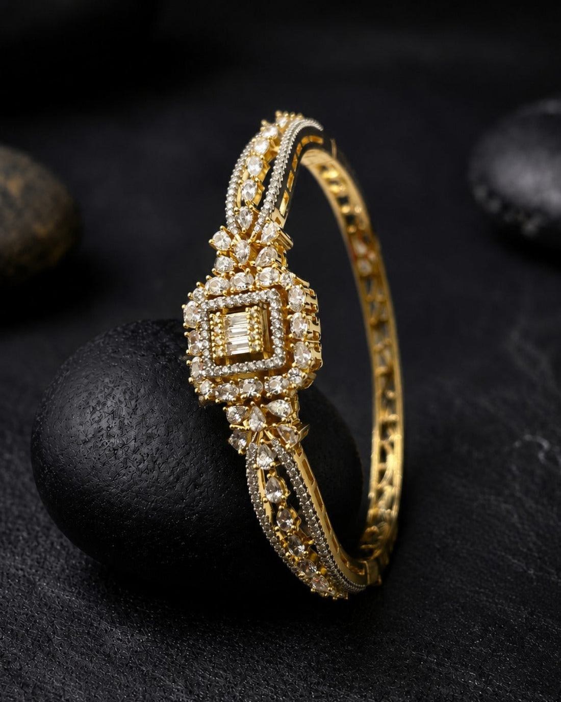 A gold hinged bangle bracelet with a square-cut diamond center and intricate pavé diamond detailing, displayed on a dark textured surface with smooth black stones.