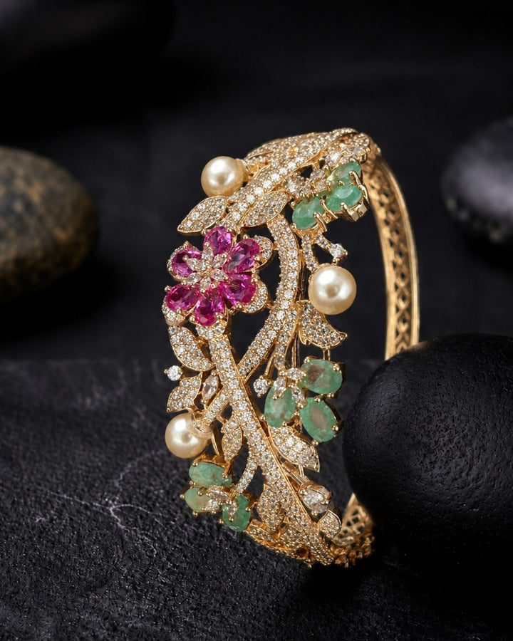 A gold hinged cuff bracelet decorated with pavé-set clear stones, green emerald-cut gemstones, white pearl beads, and a central pink gemstone floral motif, displayed on a dark textured surface with black stones.