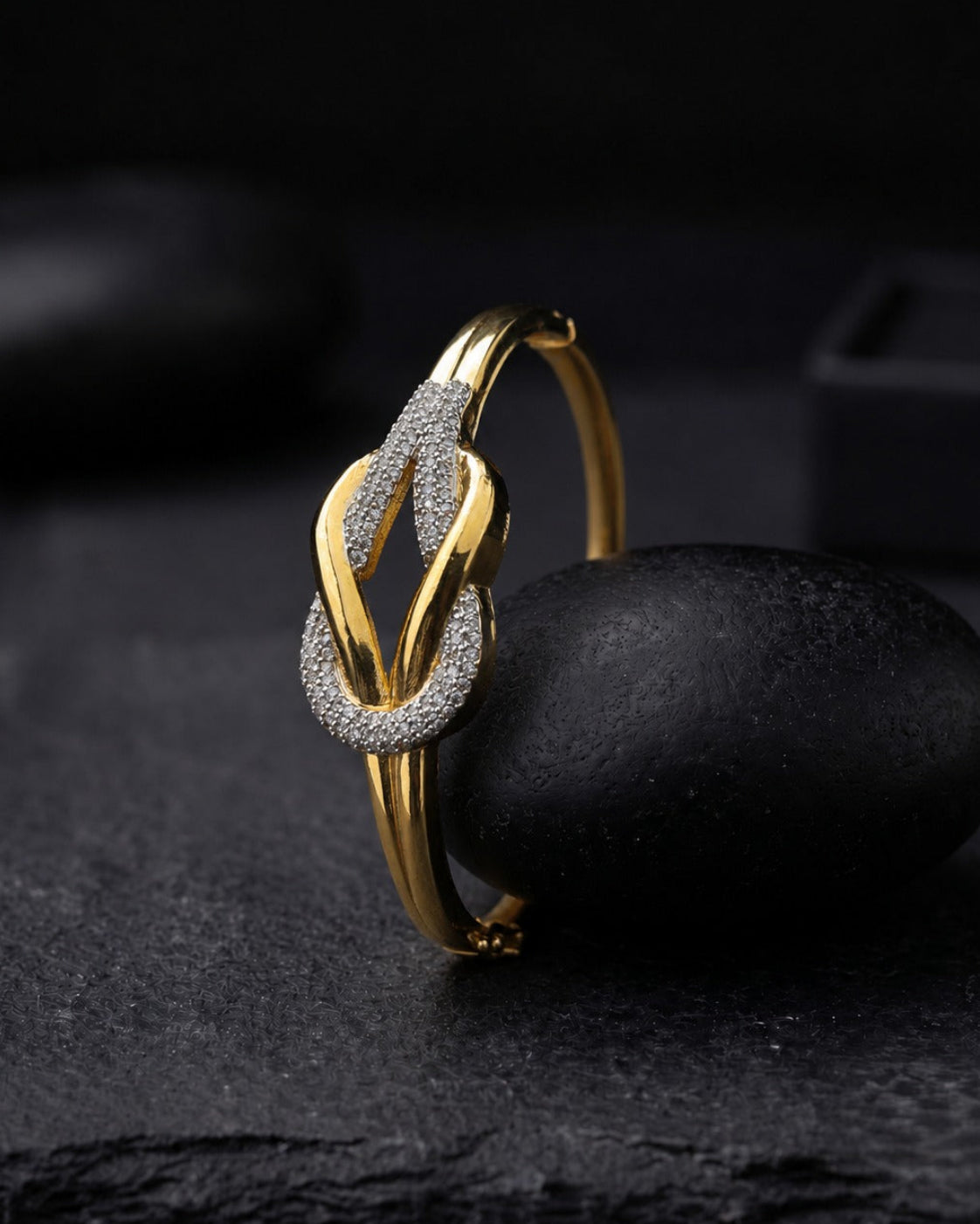 A gold hoop ring with a textured pavé-set knot design, displayed on a smooth black stone against a dark, moody background.