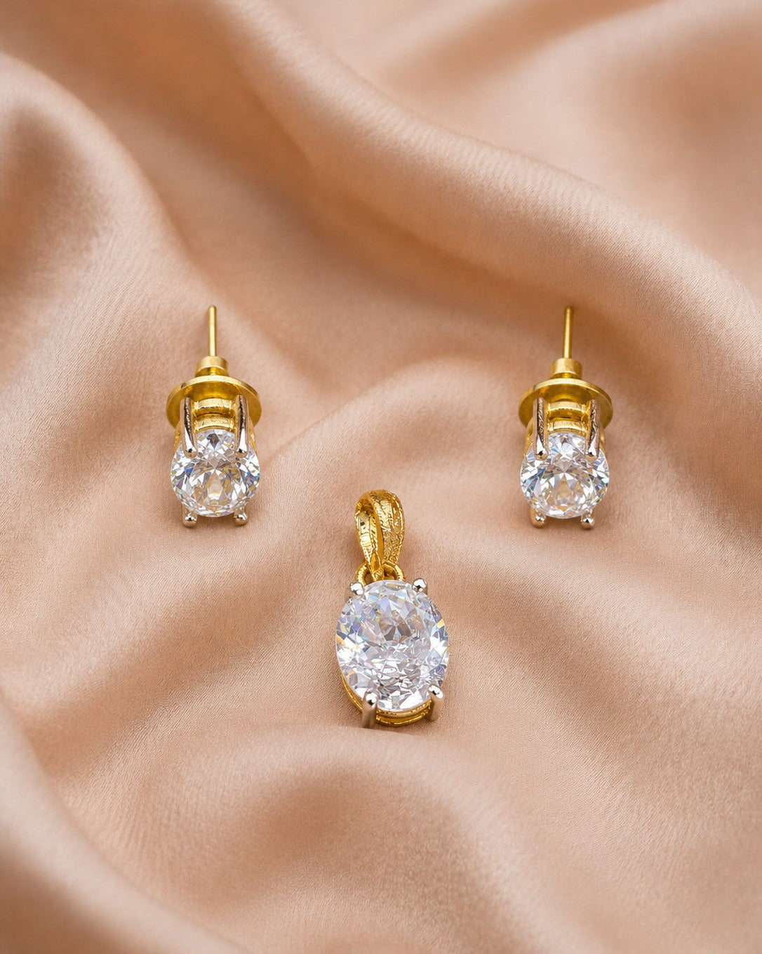 A gold jewelry set featuring a matching pendant and stud earrings with large oval clear gemstones, displayed on a draped beige satin fabric background.