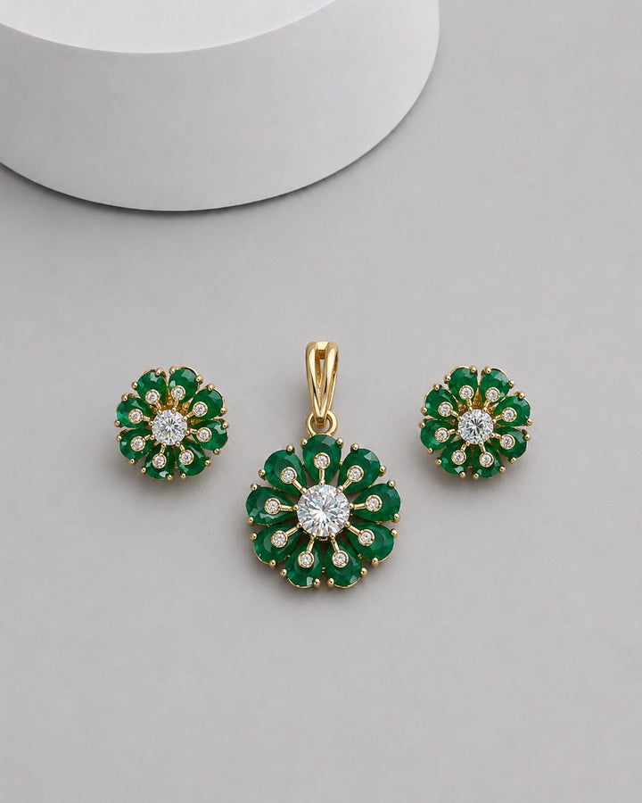 A gold jewelry set featuring a pendant and matching stud earrings, each designed as a flower with green stones and a central clear gemstone, displayed on a neutral gray surface.
