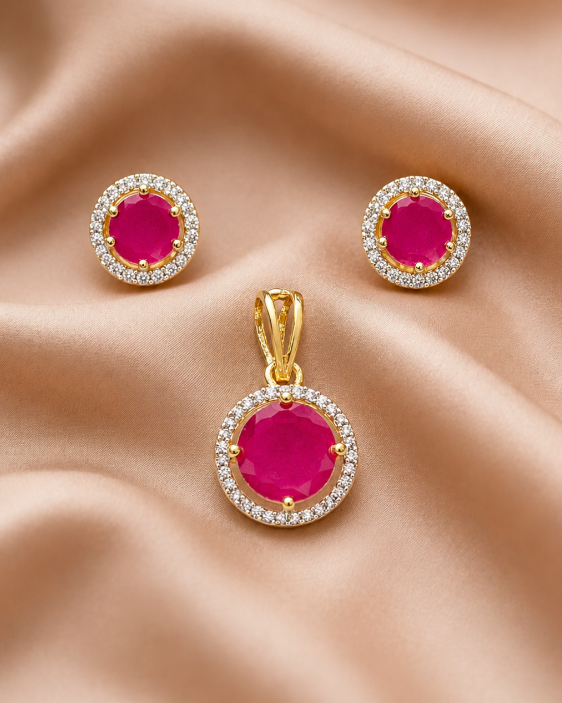 A gold jewelry set featuring a pendant and matching stud earrings, each with a round bright pink gemstone surrounded by a halo of small clear stones, displayed on a draped beige satin fabric.