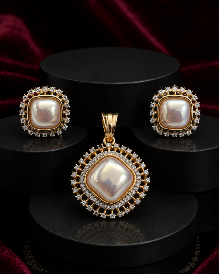 A gold jewelry set featuring a pendant, matching earrings, and a ring, all adorned with large iridescent white pearls and small clear stones, displayed on black cylindrical pedestals against a dark red velvet background.