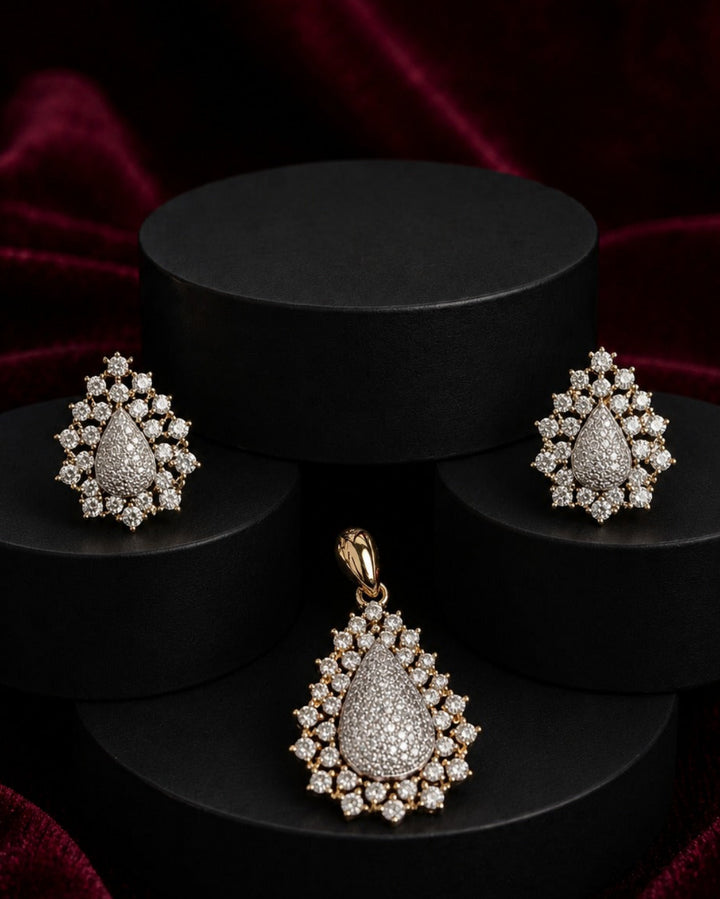 A gold jewelry set featuring a teardrop pendant and matching earrings, both adorned with sparkling clear pavé stones, displayed on a black tiered pedestal against a dark red velvet background.