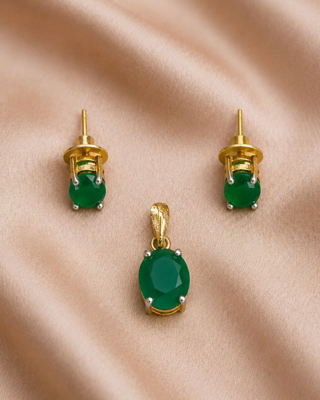 A gold jewelry set featuring an oval green stone pendant and matching stud earrings, displayed on a draped beige satin fabric background.