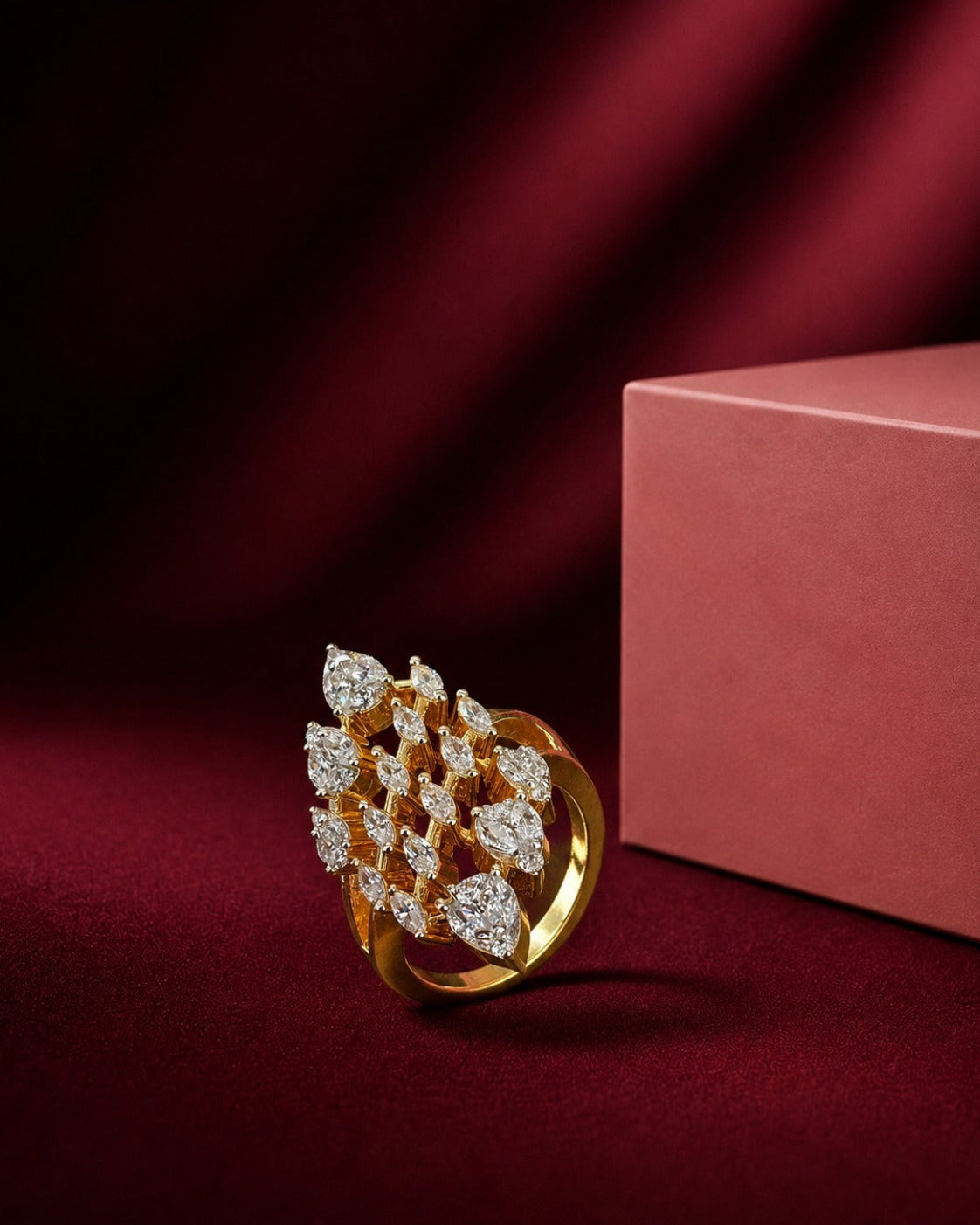 A gold ring with a cluster of marquise-cut clear gemstones, styled on a dark red velvet surface next to a dusty pink jewelry box.