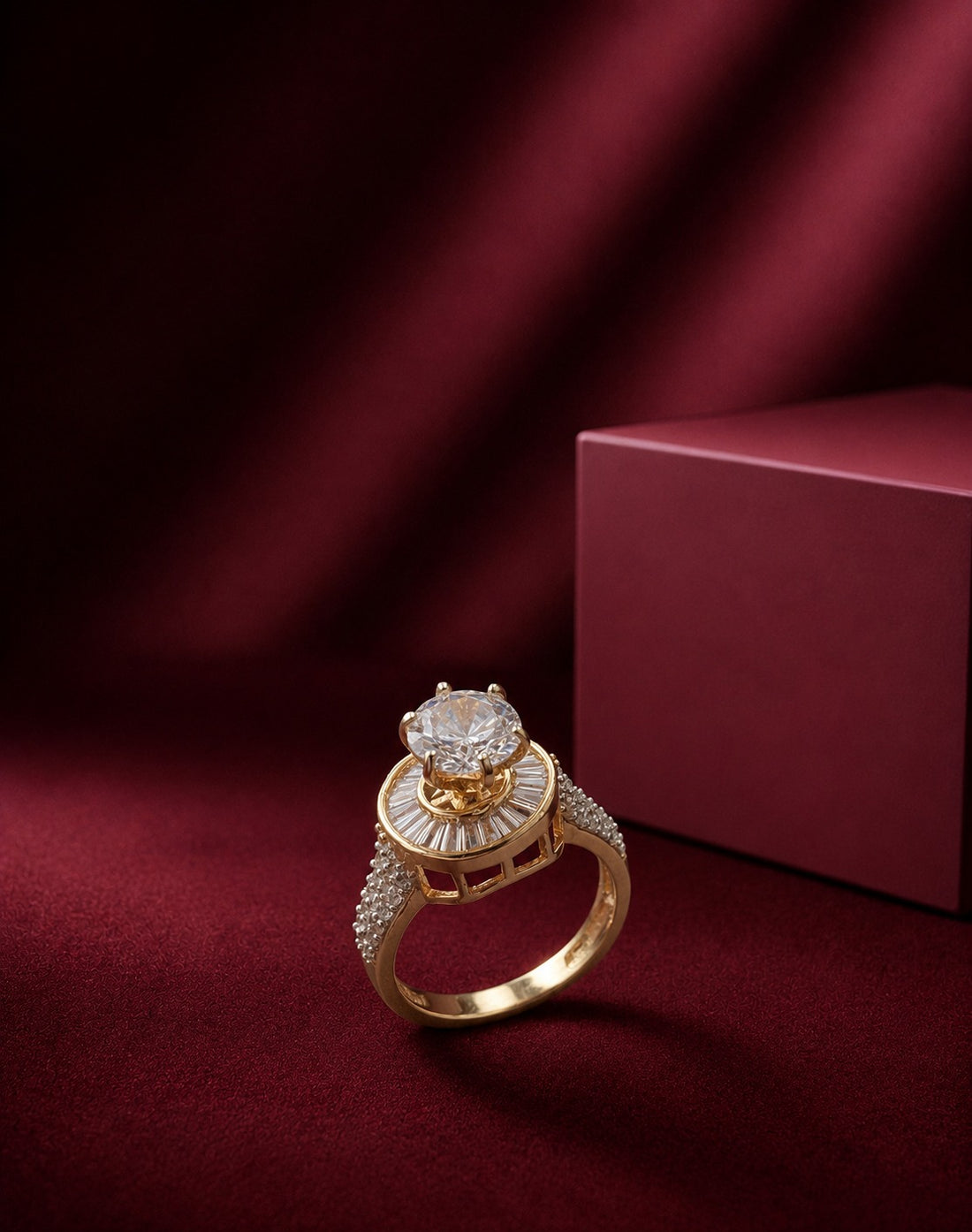 A gold ring with a large clear center stone and pavé-set side stones, displayed on a deep burgundy velvet surface next to a matching burgundy box.