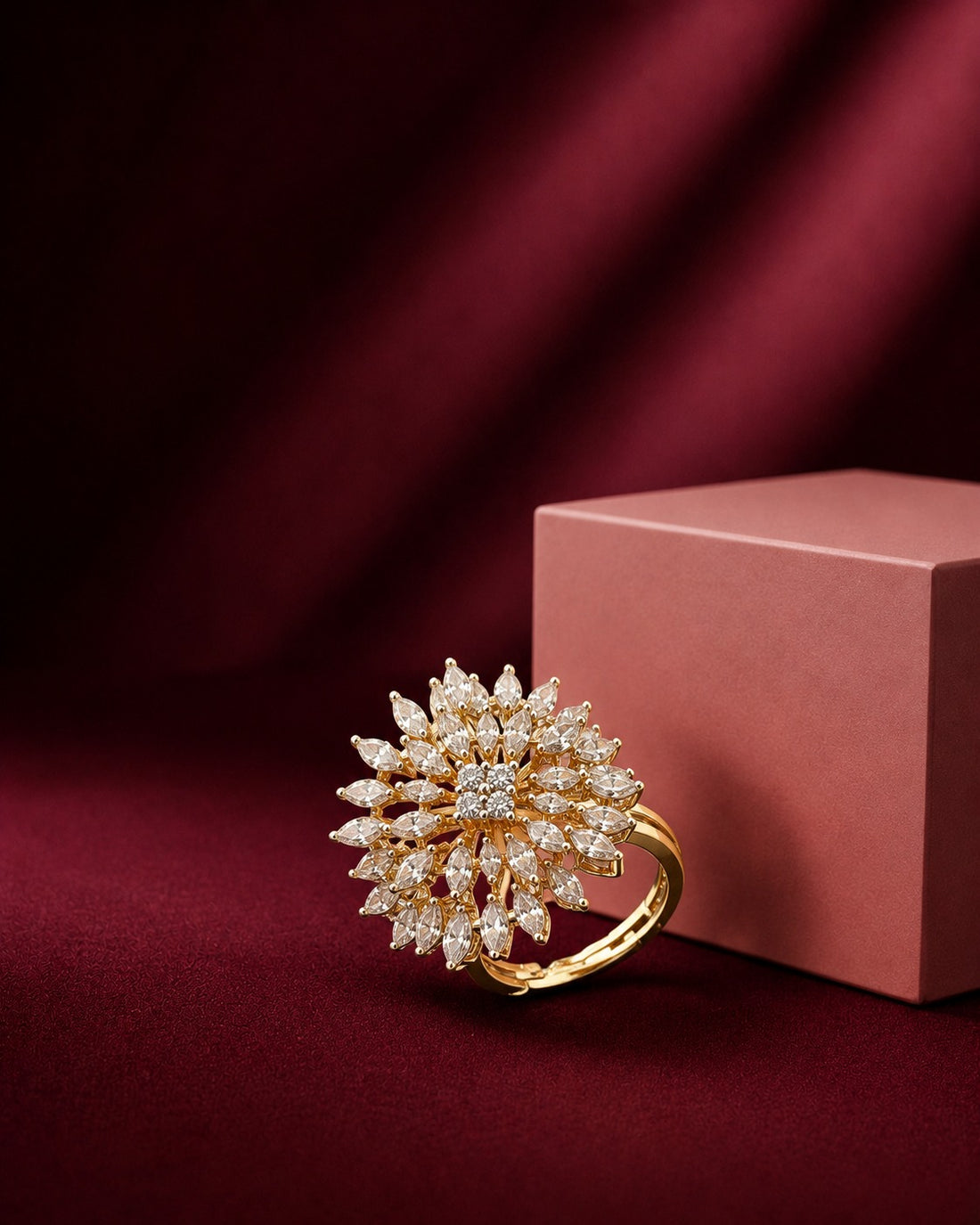 A gold ring with a large, intricate sunburst cluster of clear gemstones, displayed on a deep burgundy velvet surface next to a dusty rose jewelry box.
