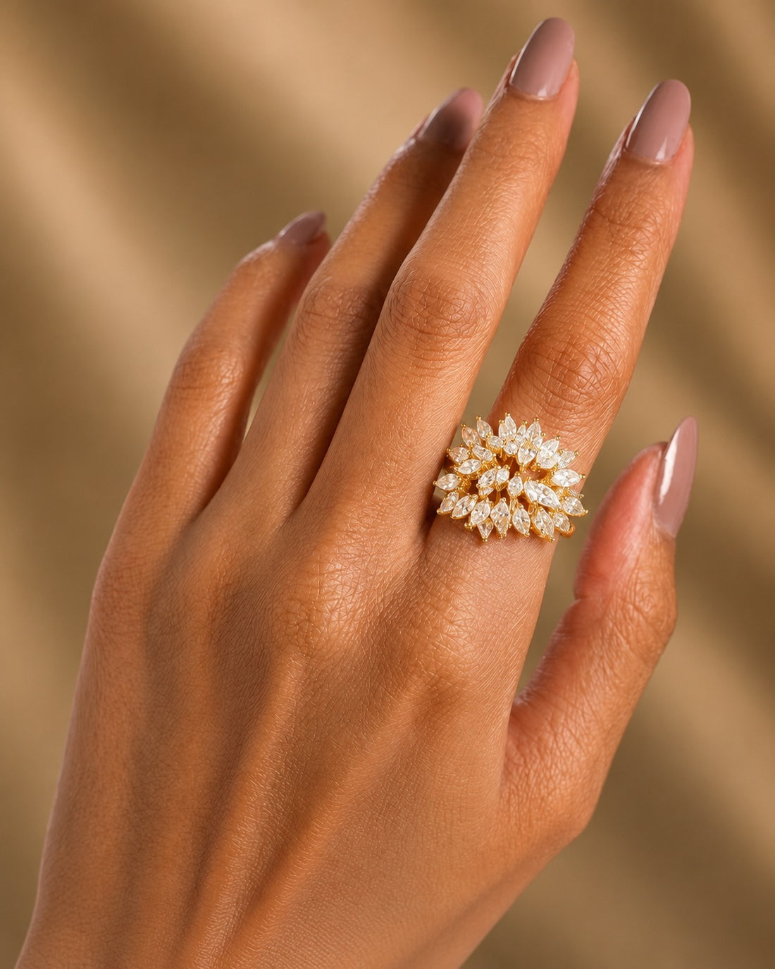 A close-up of a hand wearing a gold ring set with marquise-cut clear gemstones, set against a soft, warm beige background.