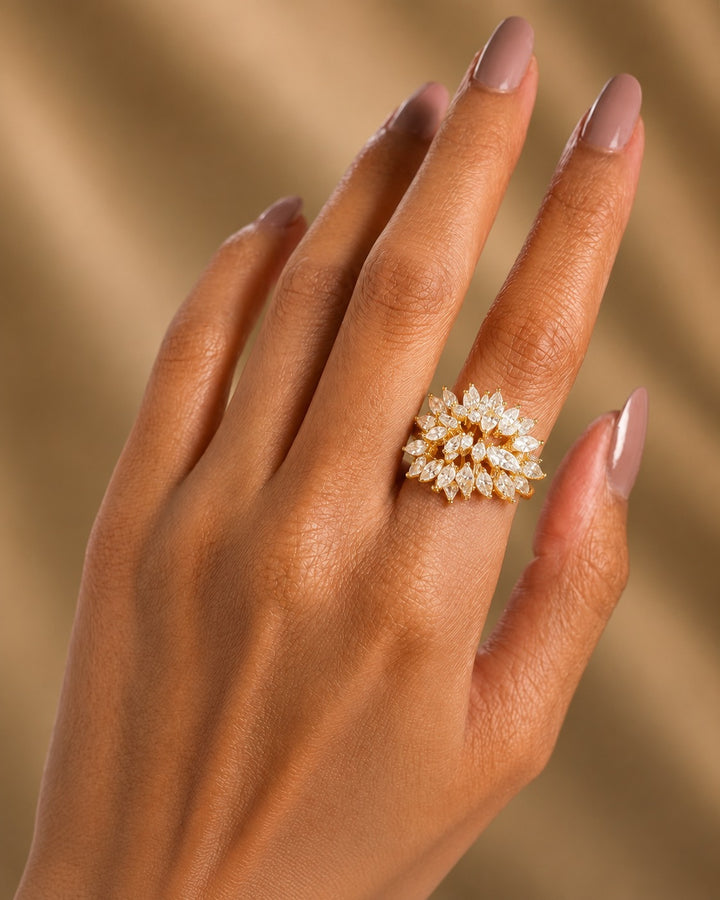A close-up of a hand wearing a gold ring set with marquise-cut clear gemstones, set against a soft, warm beige background.