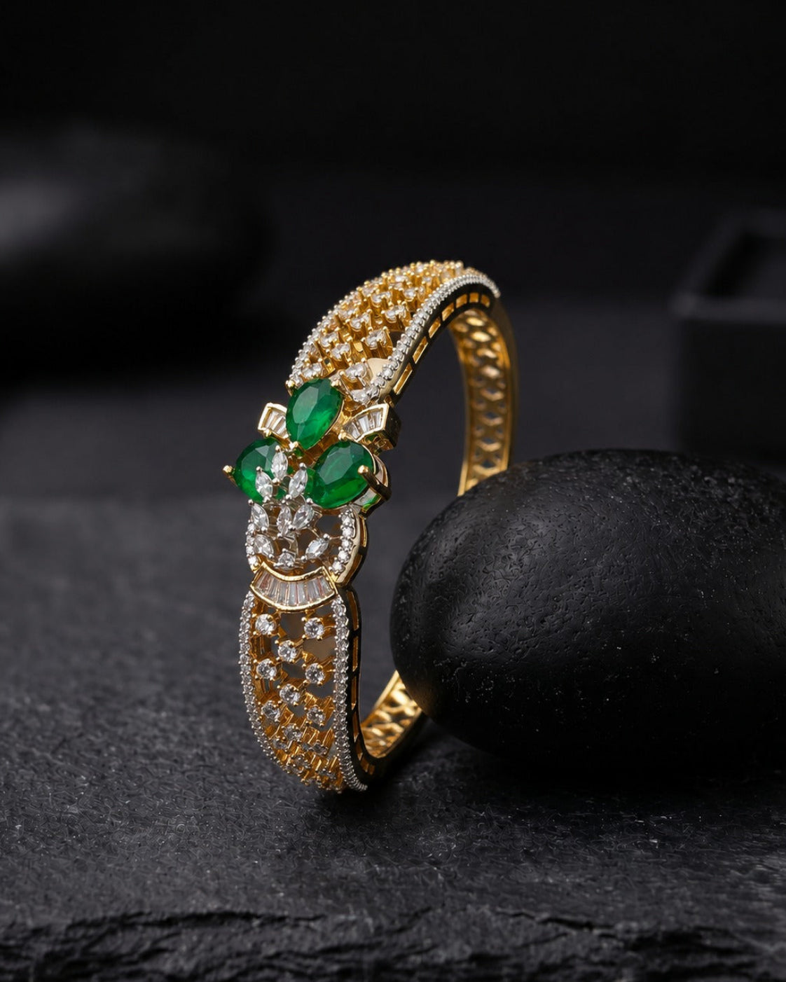 A gold ring with green emerald-cut gemstones and small clear stones, displayed on a dark textured surface next to a black stone.