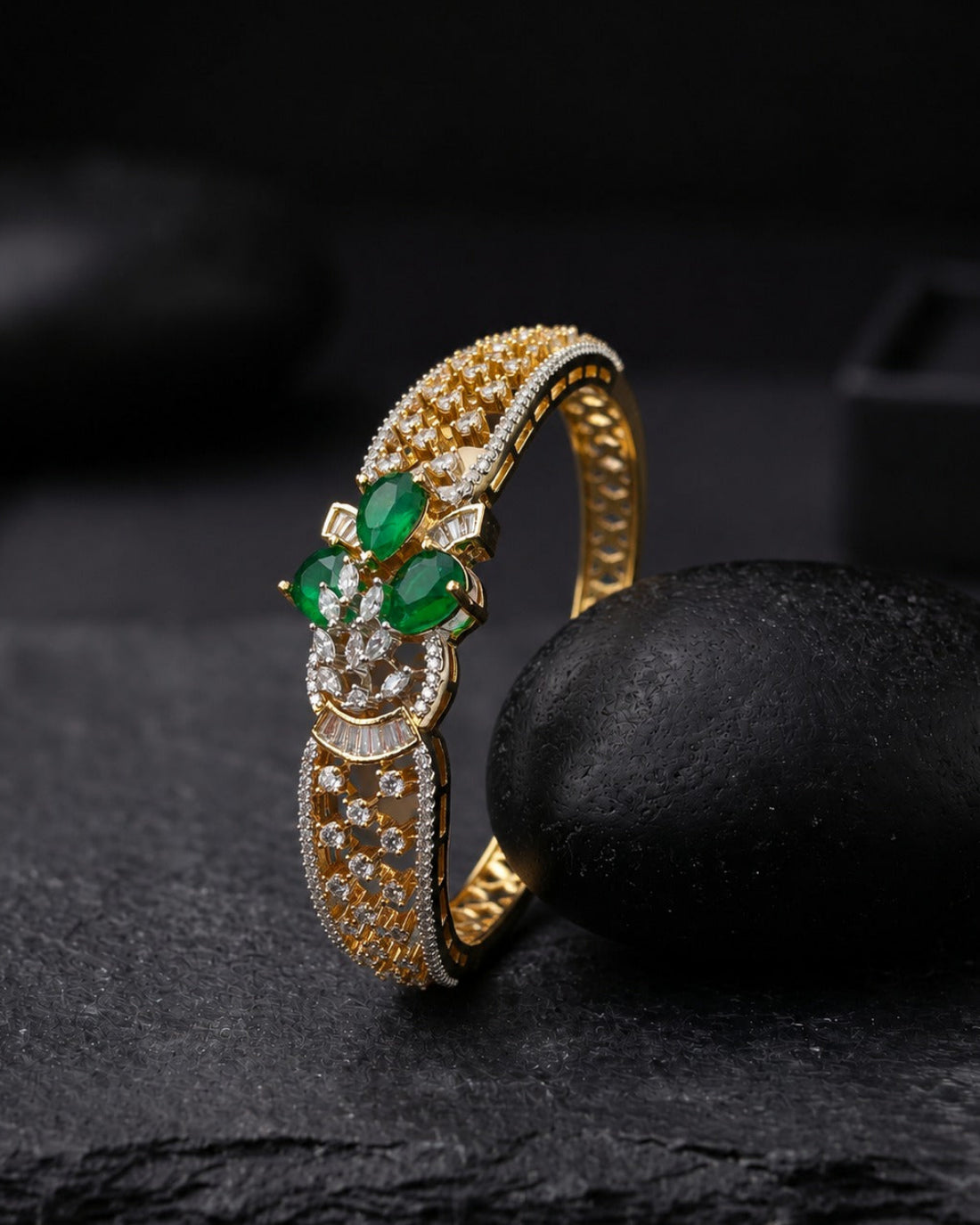 A gold ring with green emerald-cut gemstones and small clear stones, displayed on a dark textured surface next to a black stone.