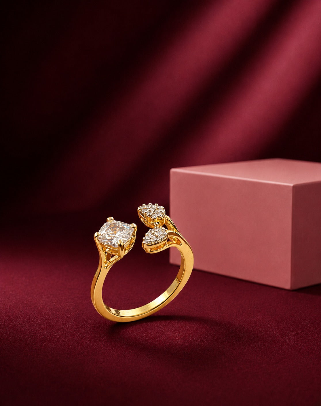 A gold ring with two diamond-set floral motifs, displayed on a dark red fabric surface next to a pink box.