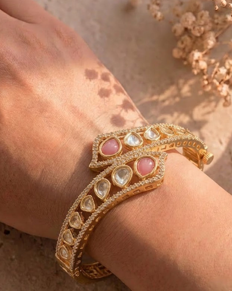 A close-up of a person's wrist wearing a gold-toned traditional Indian style bracelet adorned with pink and clear stones, set against a warm beige textured background with dried flowers.