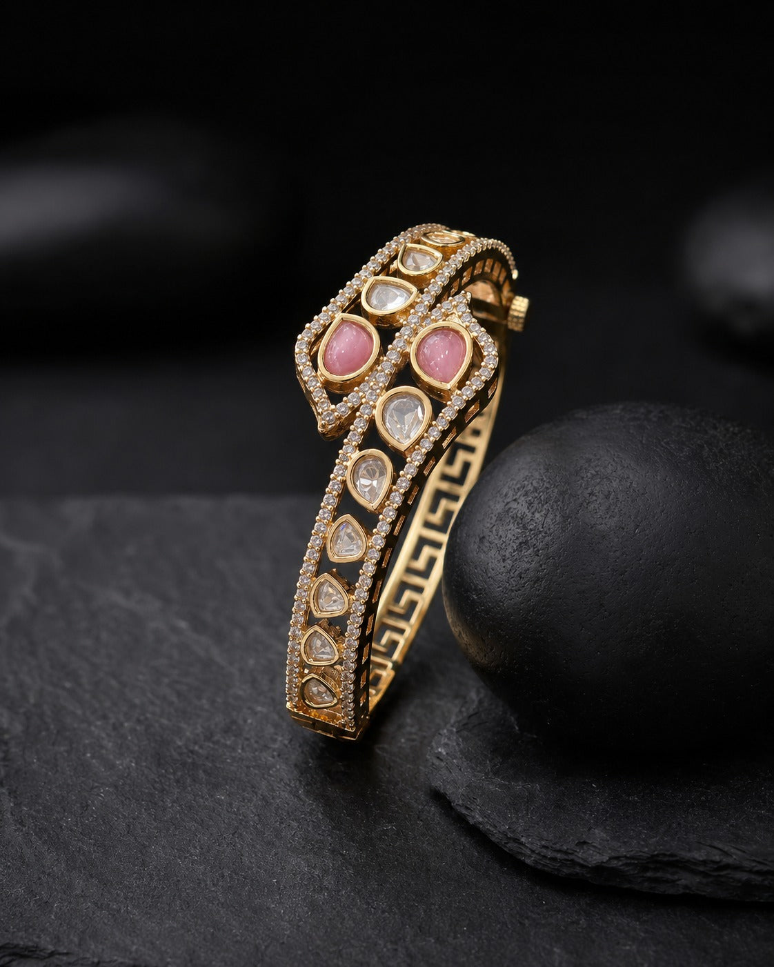A gold-plated snake-head cuff bracelet with pink and clear gemstone accents, displayed on a dark slate surface.