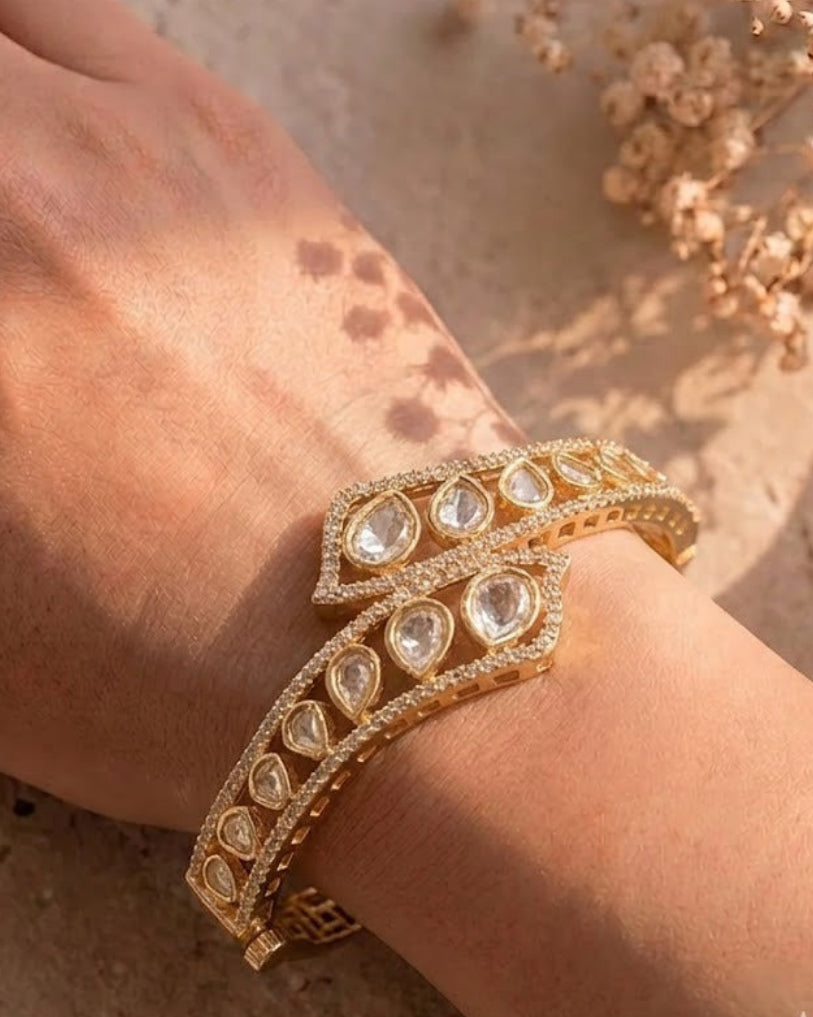 A gold-toned double bangle bracelet with clear teardrop-shaped stones and pavé detailing, shown worn on a person's wrist against a warm beige textured background with dried flowers.