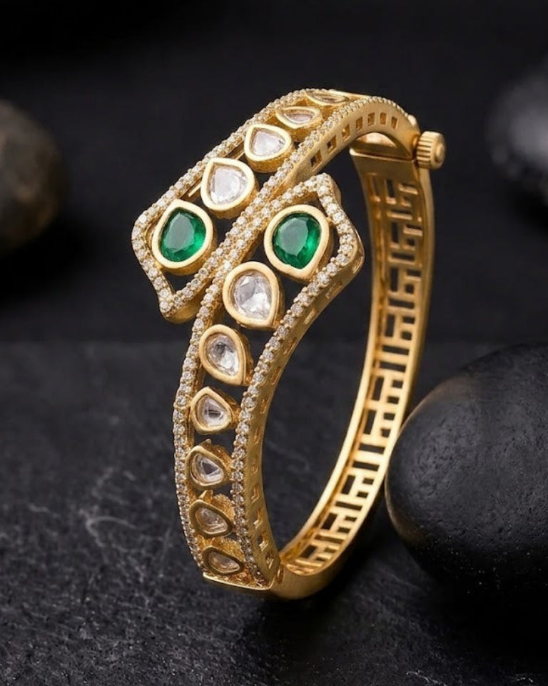 A gold-plated snake-shaped cuff bracelet with green and clear gemstone accents, displayed on a dark textured surface with black stones.