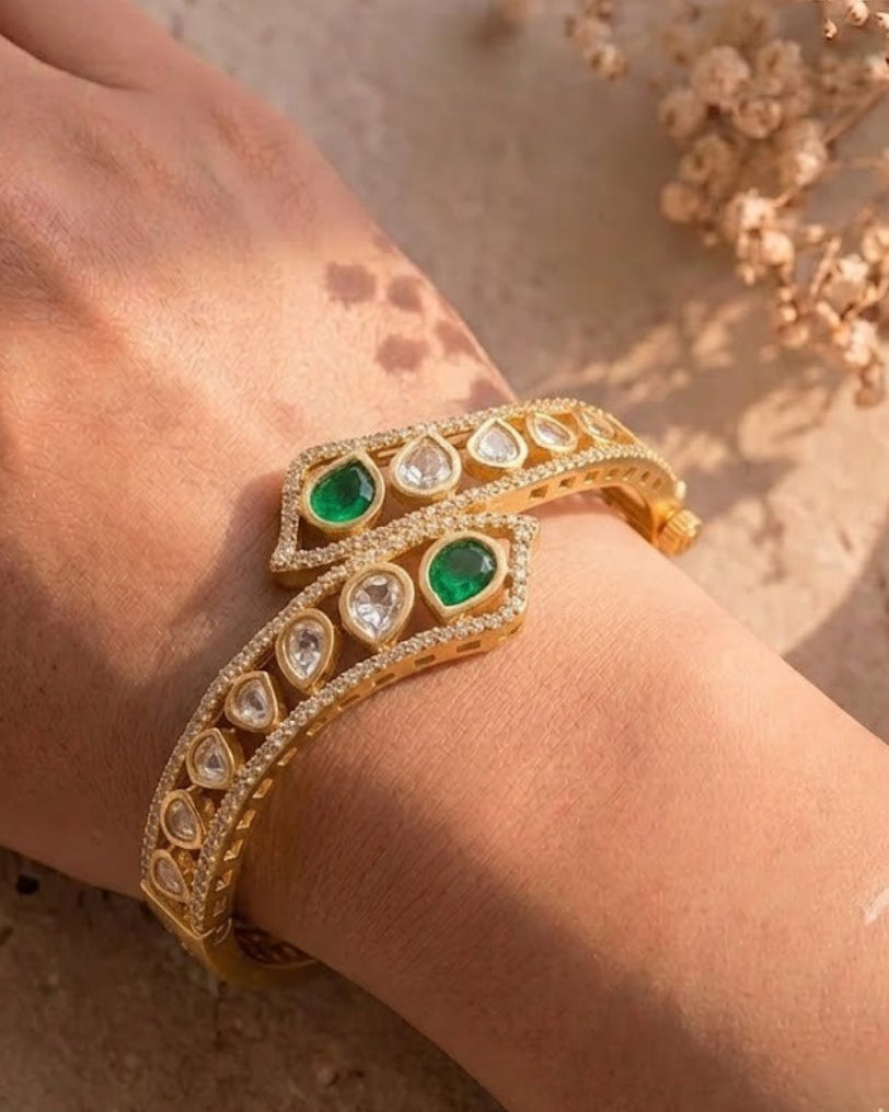A gold-plated double bangle bracelet with green emerald-cut stones and clear pavé-set stones, shown worn on a person's wrist against a warm beige textured background with dried flowers.