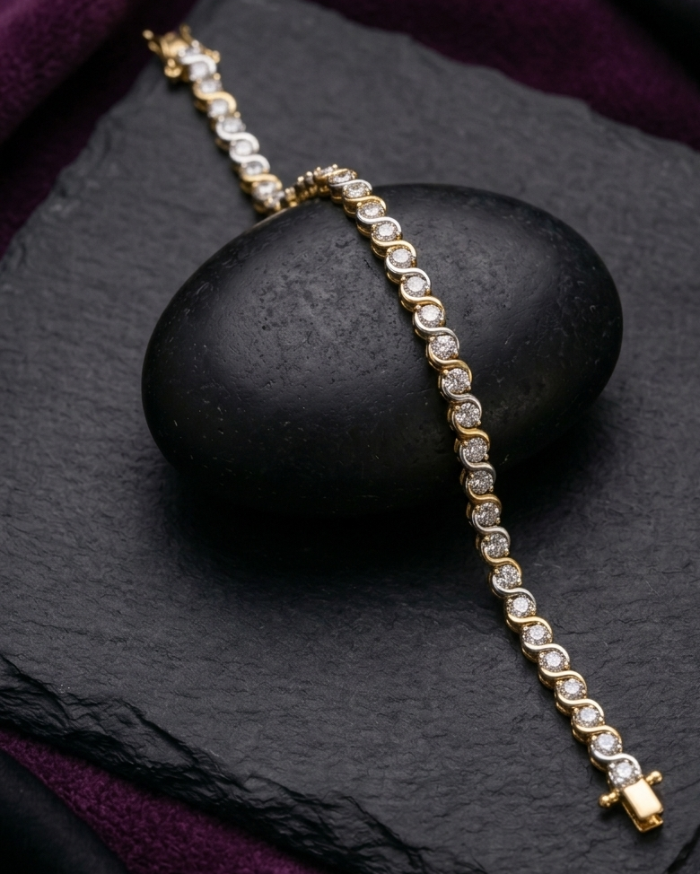 A gold tennis bracelet with clear round-cut stones, elegantly draped over a smooth black stone on a dark slate surface with deep purple fabric in the background.