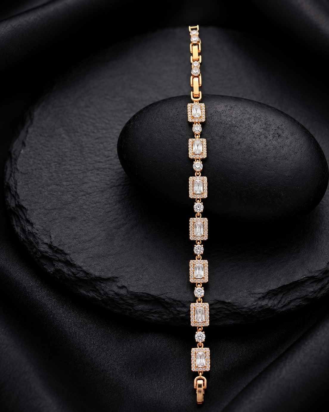 A gold tennis bracelet with rectangular emerald-cut clear stones, displayed vertically on a black stone stand against a black fabric background.