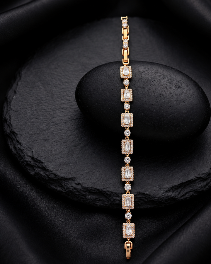 A gold tennis bracelet with rectangular emerald-cut clear stones, displayed vertically on a black stone stand against a black fabric background.