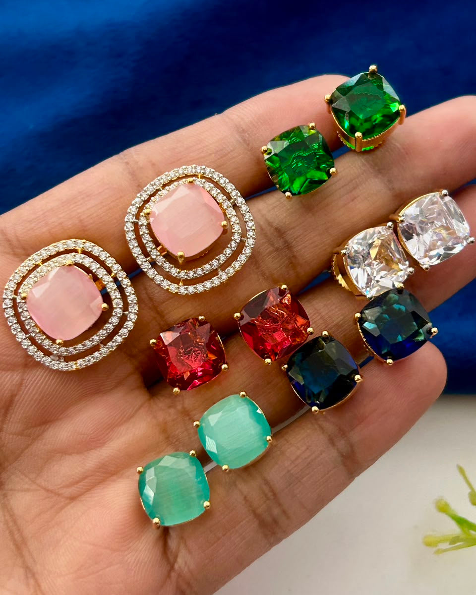 A hand holding multiple pairs of stud earrings with colorful gemstones in gold settings, including pink, green, red, blue, and clear stones, against a blue fabric background.