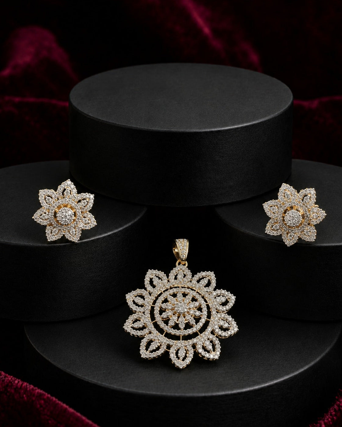 A jewelry set featuring a floral pendant and matching stud earrings in a gold-tone setting with clear pavé stones, displayed on black cylindrical pedestals against a dark red velvet background.