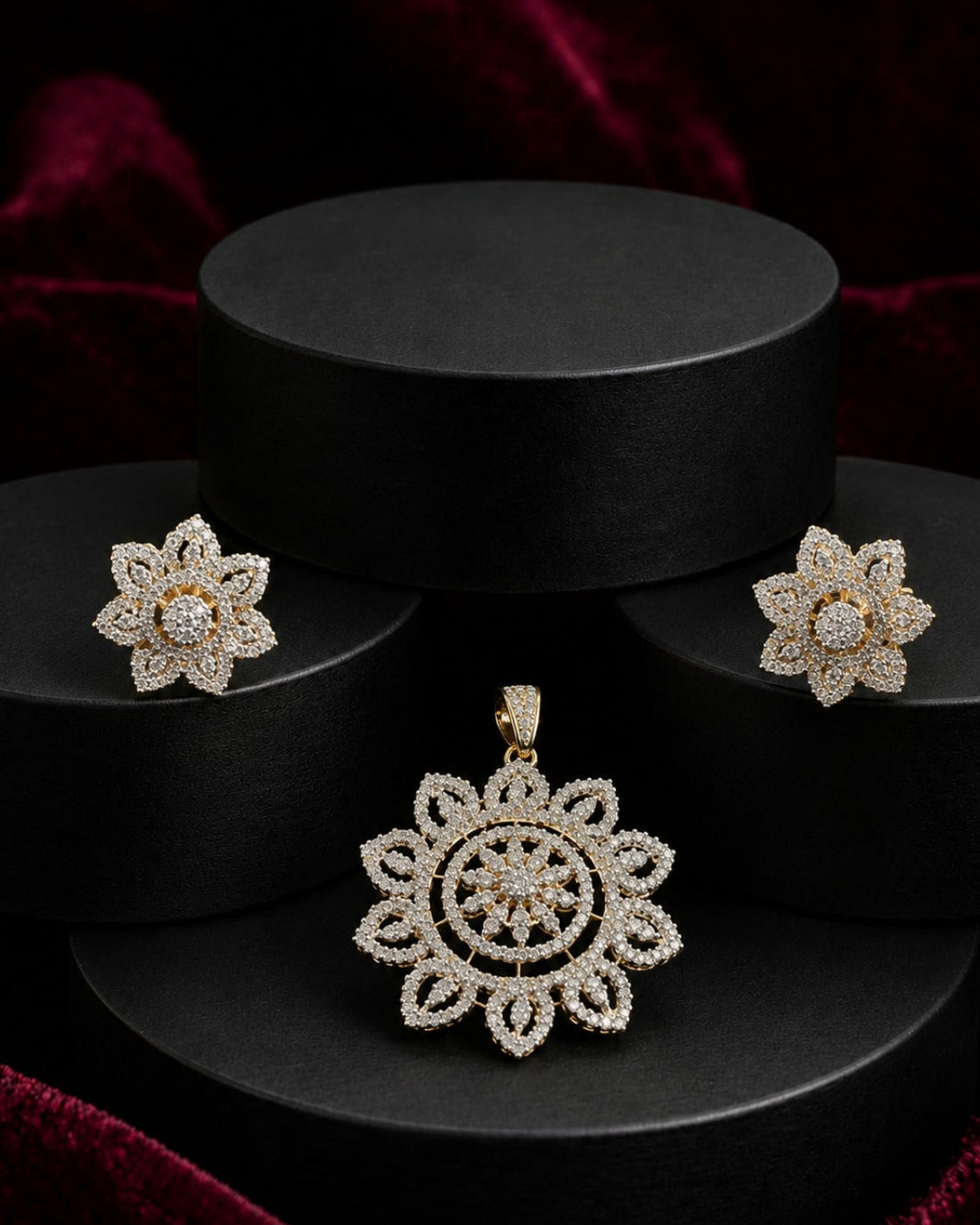 A jewelry set featuring a floral pendant and matching stud earrings in a gold-tone setting with clear pavé stones, displayed on black cylindrical pedestals against a dark red velvet background.