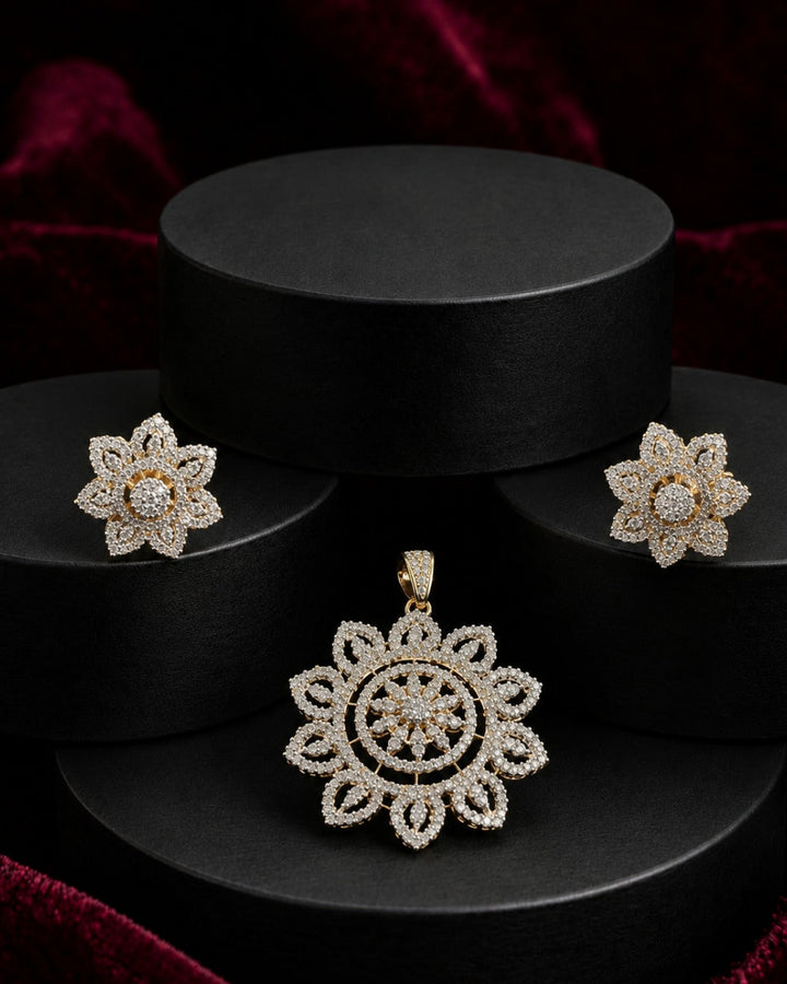 A jewelry set featuring a floral pendant and matching stud earrings in a gold-tone setting with clear pavé stones, displayed on black cylindrical pedestals against a dark red velvet background.