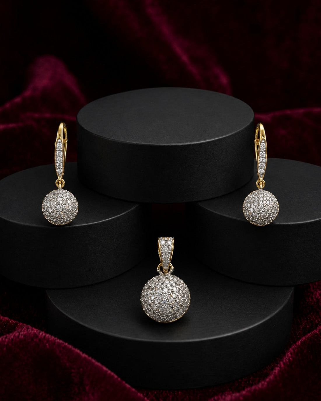 A jewelry set featuring a gold pendant and matching drop earrings, both adorned with pavé-set clear stones, displayed on a black tiered pedestal against a dark red velvet background.