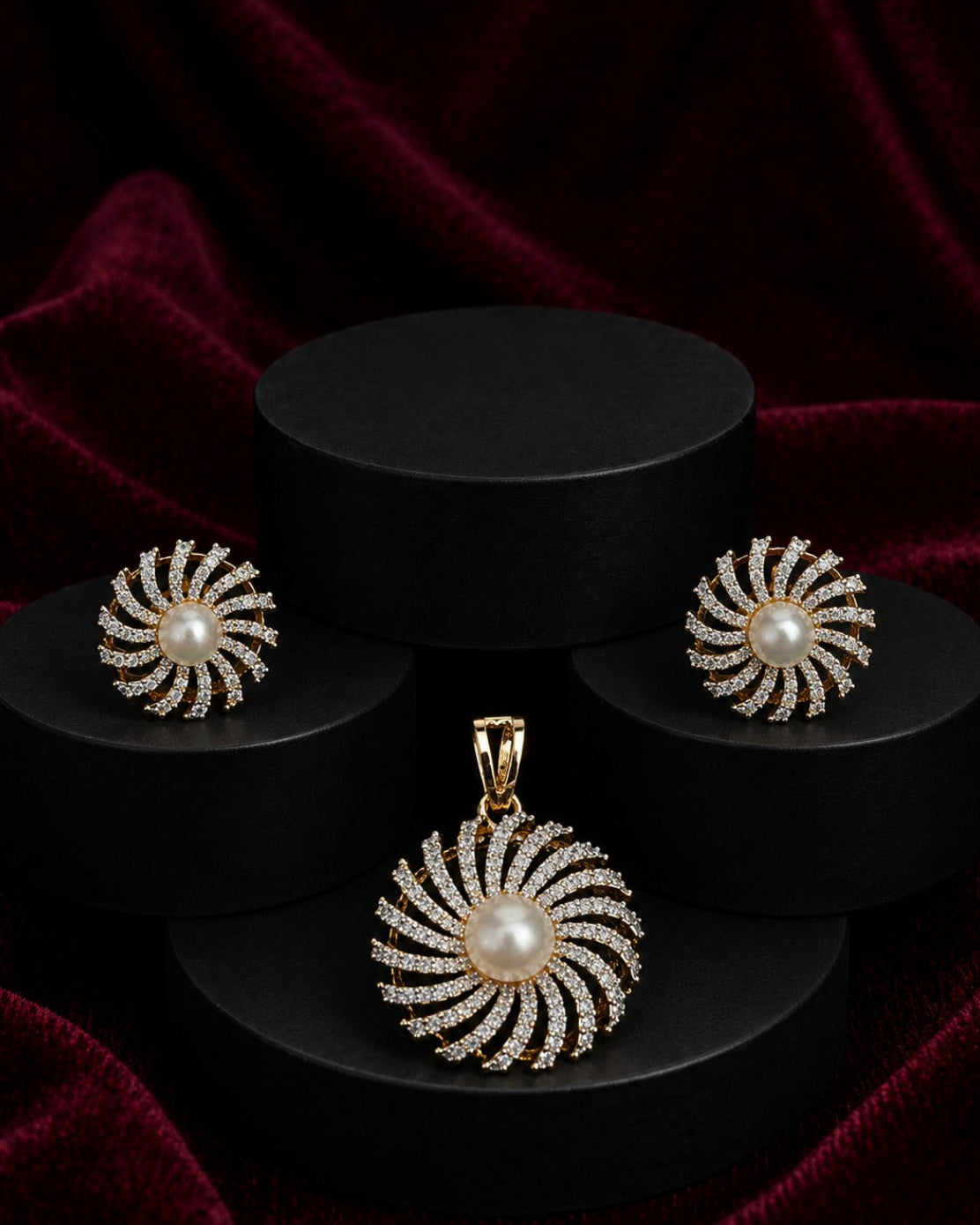 A jewelry set featuring a gold pendant and matching earrings with white pearl centers and surrounding clear stones, arranged on black cylindrical pedestals against a dark red velvet background.