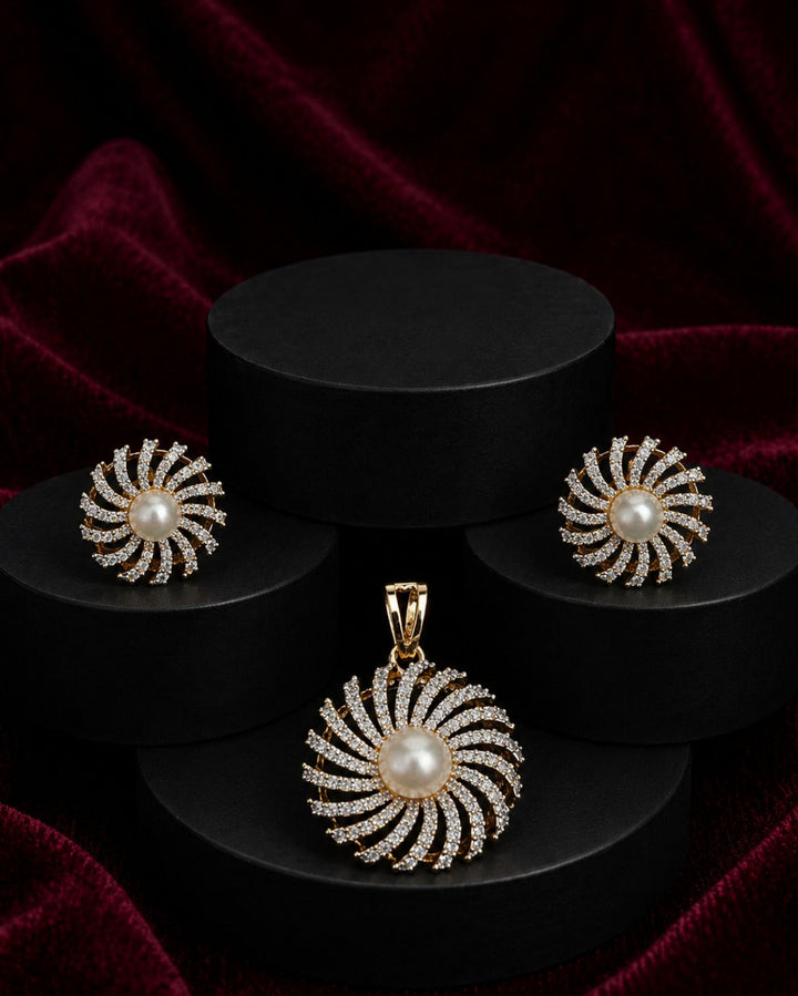 A jewelry set featuring a gold pendant and matching earrings with white pearl centers and surrounding clear stones, arranged on black cylindrical pedestals against a dark red velvet background.