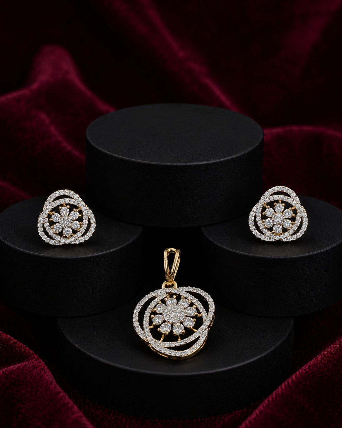 A jewelry set featuring a gold pendant and matching stud earrings with intricate circular floral designs set with clear stones, displayed on black cylindrical pedestals against a dark red velvet background.