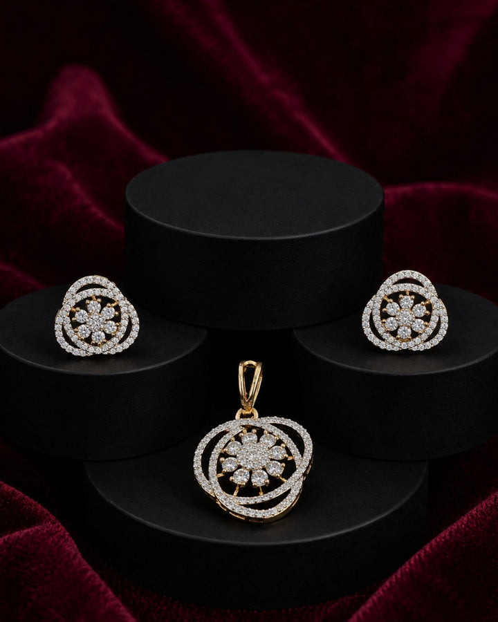 A jewelry set featuring a gold pendant and matching stud earrings with intricate circular floral designs set with clear stones, displayed on black cylindrical pedestals against a dark red velvet background.