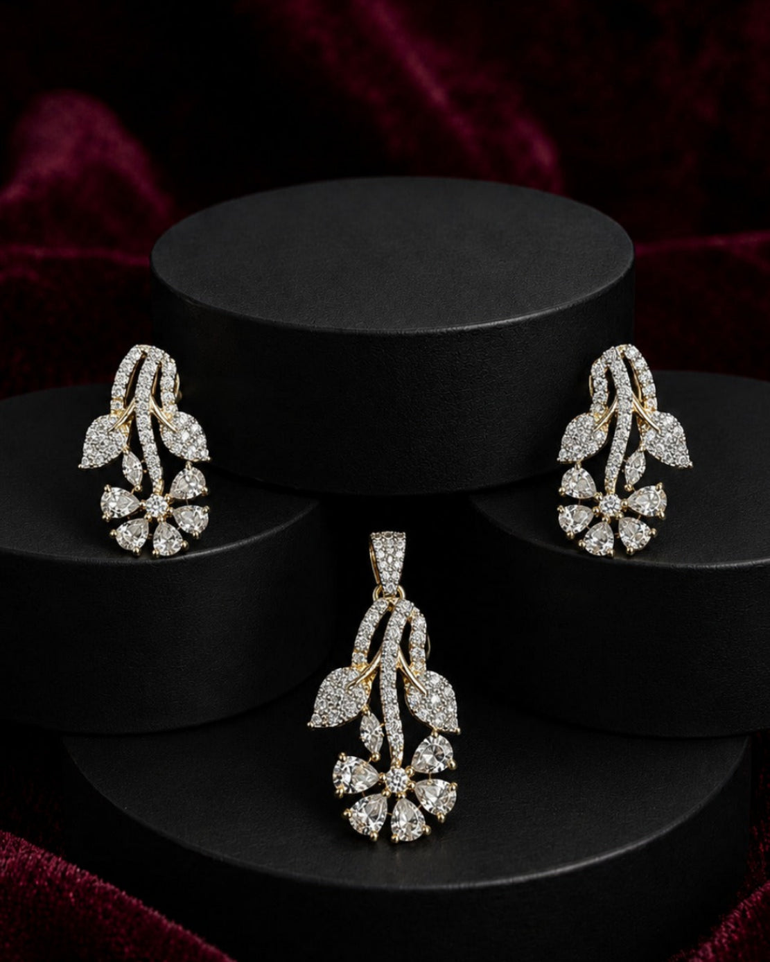 A jewelry set featuring a matching pendant and earring in yellow gold and clear stones, displayed on black cylindrical pedestals against a dark red velvet background.
