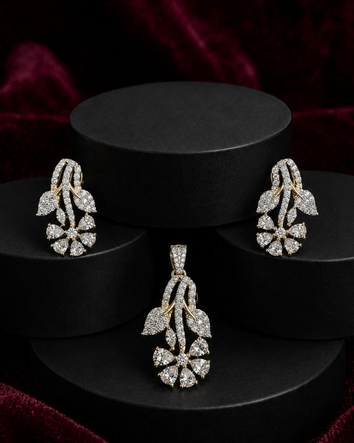 A jewelry set featuring a matching pendant and earring in yellow gold and clear stones, displayed on black cylindrical pedestals against a dark red velvet background.