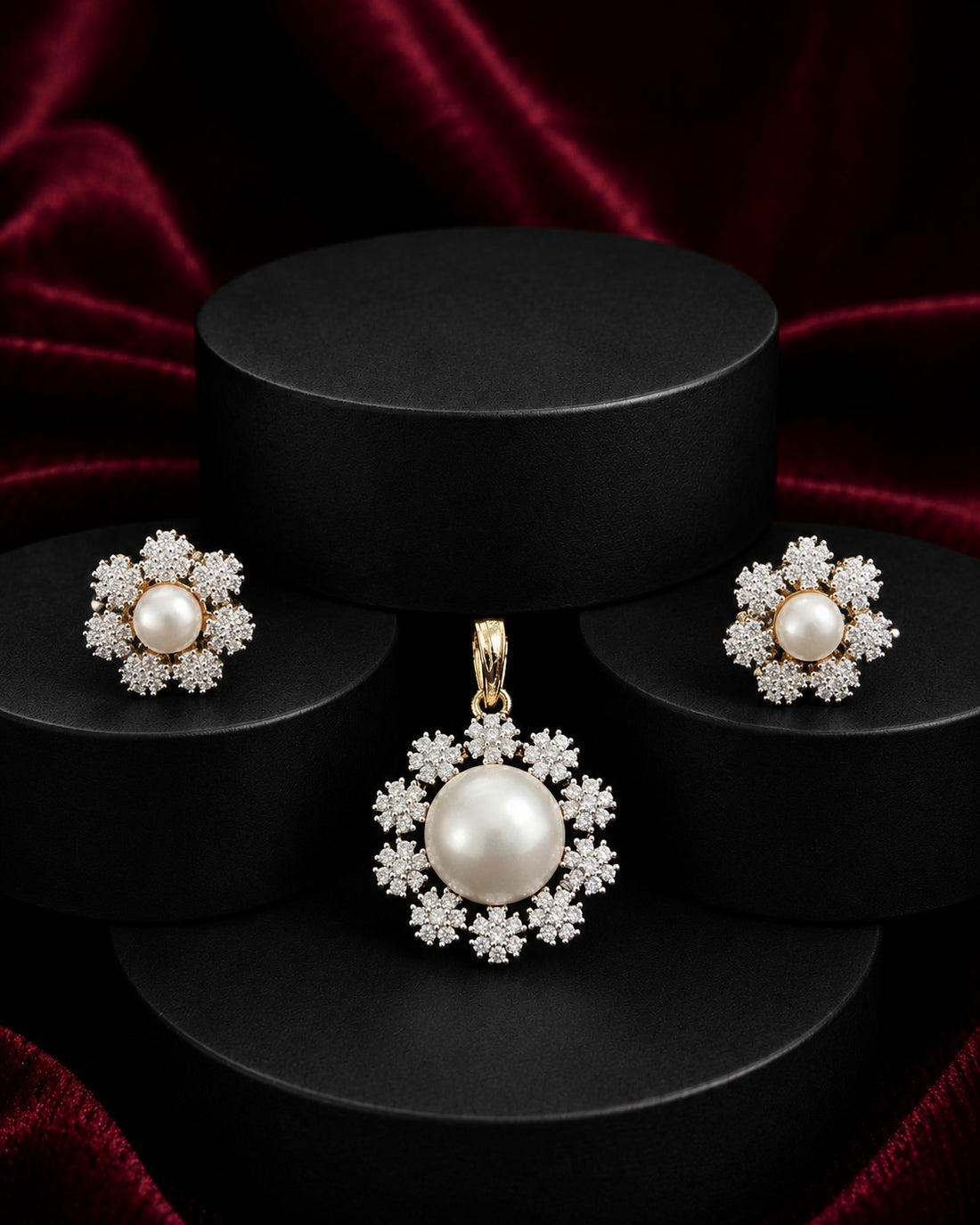 A jewelry set featuring a pearl pendant, matching earrings, and a ring, all with gold-toned settings and clear crystal-like accents, displayed on black cylindrical pedestals against a dark red velvet background.