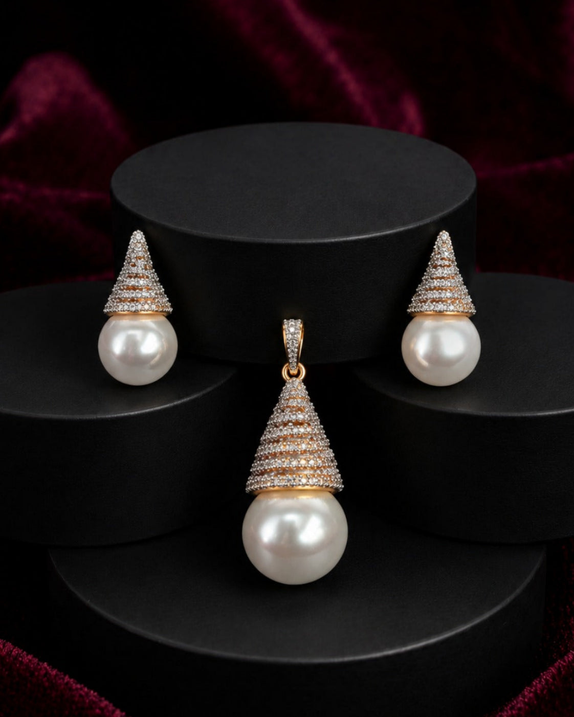 A jewelry set featuring a pendant and matching earrings with large white pearls and gold-toned settings accented with small clear stones, displayed on black cylindrical pedestals against a dark red velvet background.