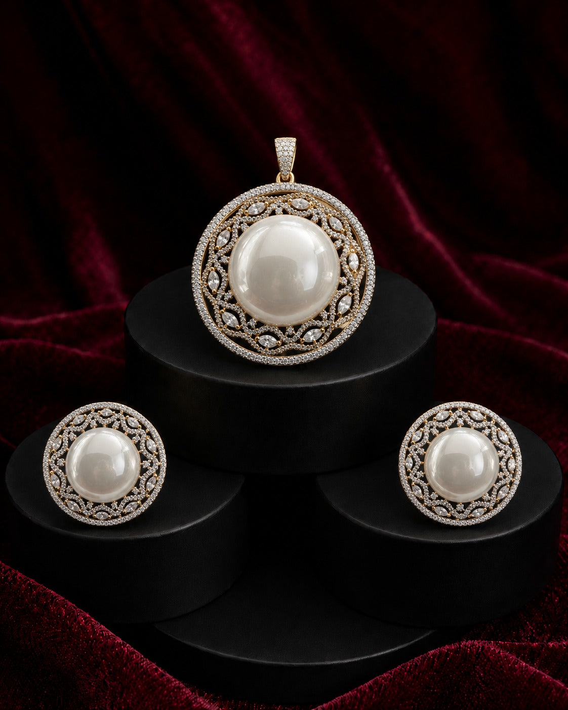A jewelry set featuring a round pendant and two matching earrings with large white pearls and ornate silver filigree borders, displayed on black cylindrical pedestals against a dark red velvet background.