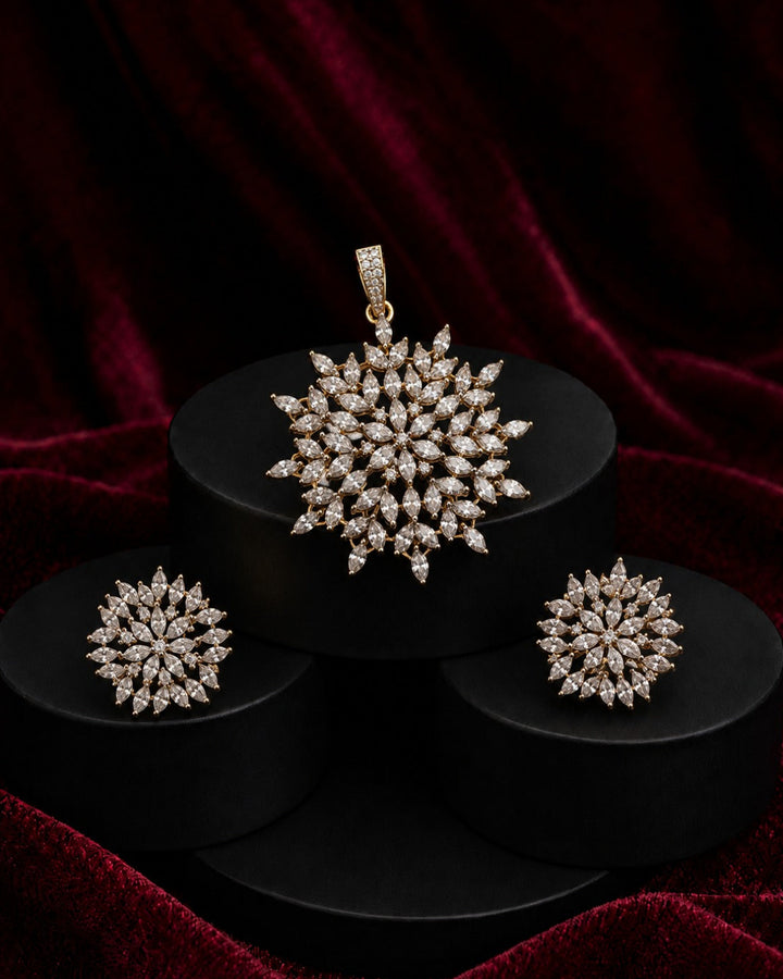 A jewelry set featuring a snowflake-shaped pendant, matching earrings, and a ring, all set in yellow gold and adorned with clear stones, displayed on black pedestals against a dark red velvet background.