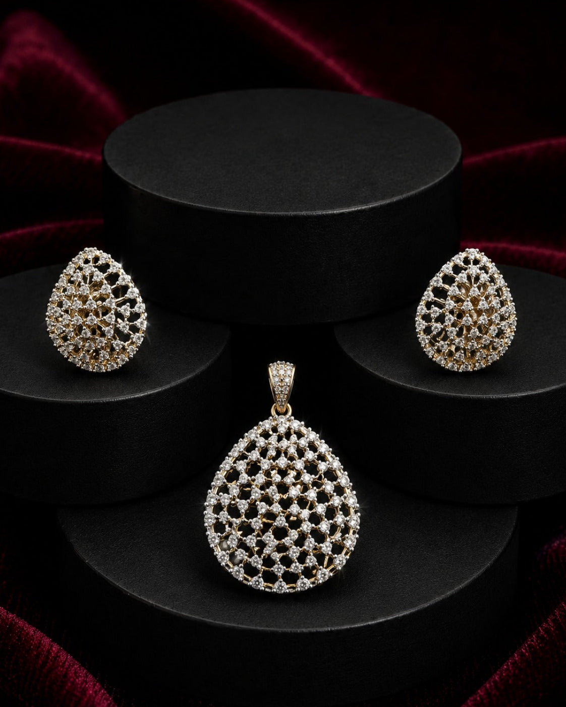 A jewelry set featuring a teardrop-shaped pendant and matching stud earrings, both adorned with intricate pavé-style diamond-like detailing in a gold-tone setting, displayed on black cylindrical pedestals against a dark red velvet backdrop.