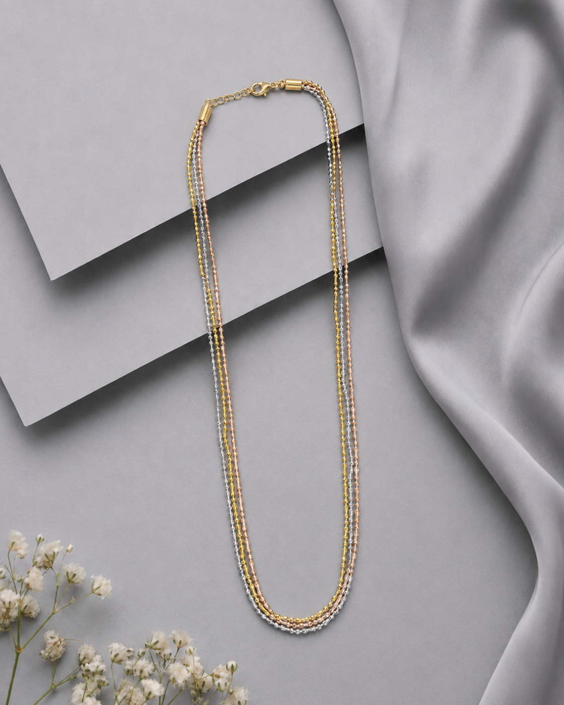 A multi-strand beaded necklace in gold and silver tones, styled on a gray background with layered geometric blocks, draped satin fabric, and small white flowers.