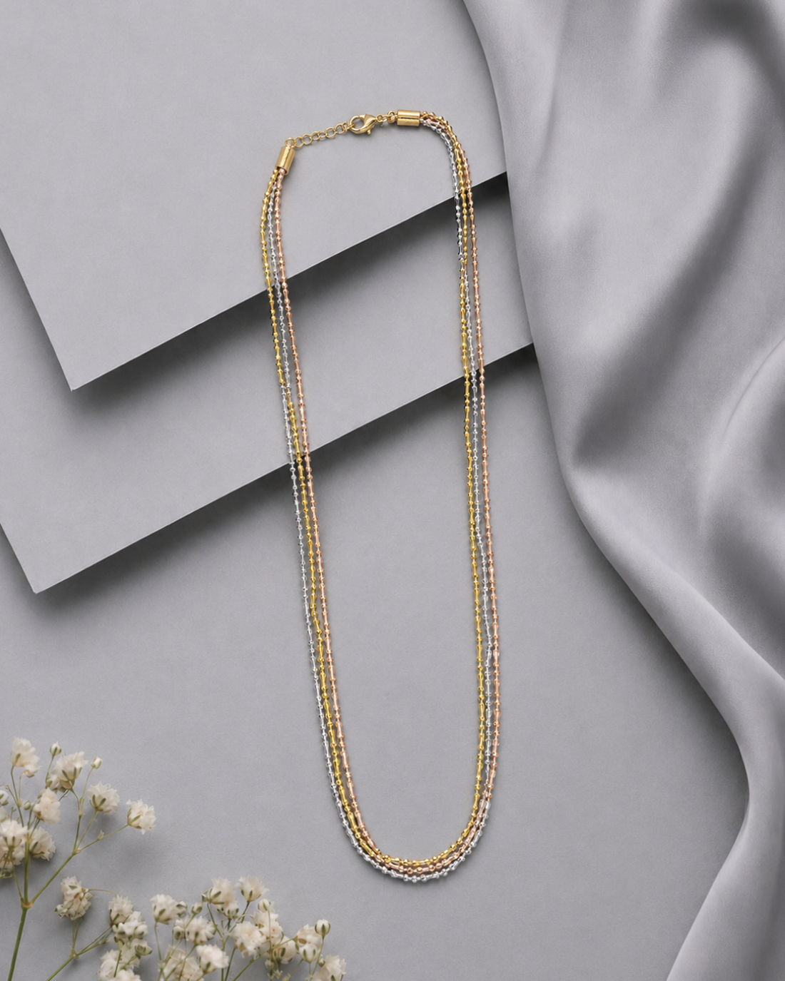 A multi-strand beaded necklace in gold and silver tones, styled on a gray background with layered geometric blocks, draped satin fabric, and small white flowers.