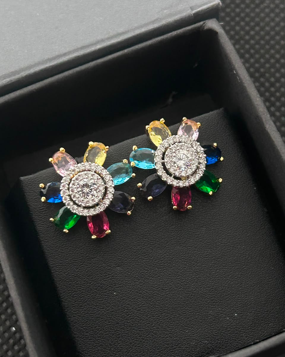 A pair of colorful flower-shaped stud earrings displayed on a black velvet jewelry cushion inside a black gift box. The earrings feature a gold-tone circular setting with clear stones, surrounded by multicolored gemstone petals in pink, yellow, blue, green, and red.