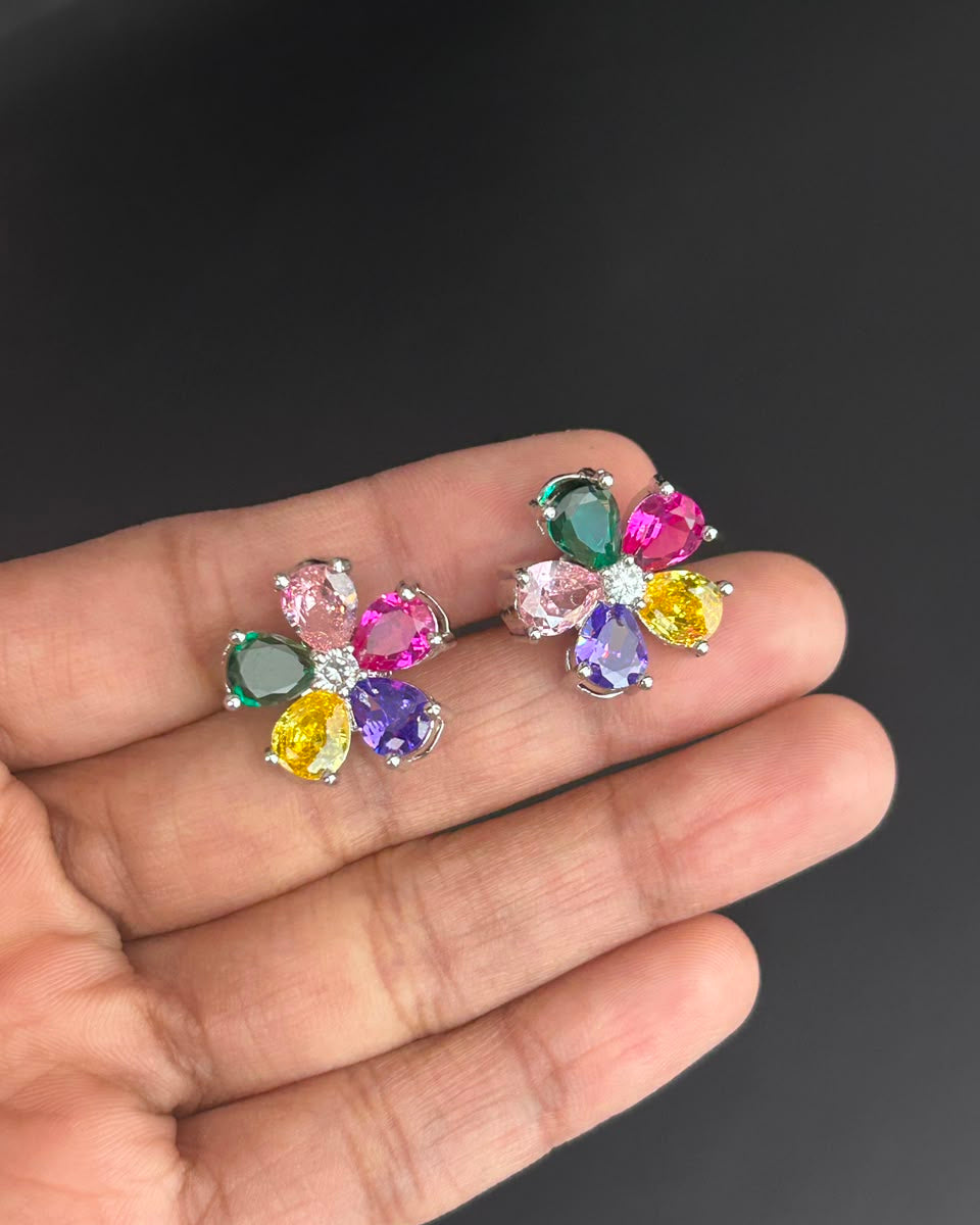 A pair of colorful flower-shaped stud earrings held on a person's hand against a dark gray background. Each earring has multiple faceted gemstones in green, pink, purple, yellow, and clear, set in a silver-tone metal.