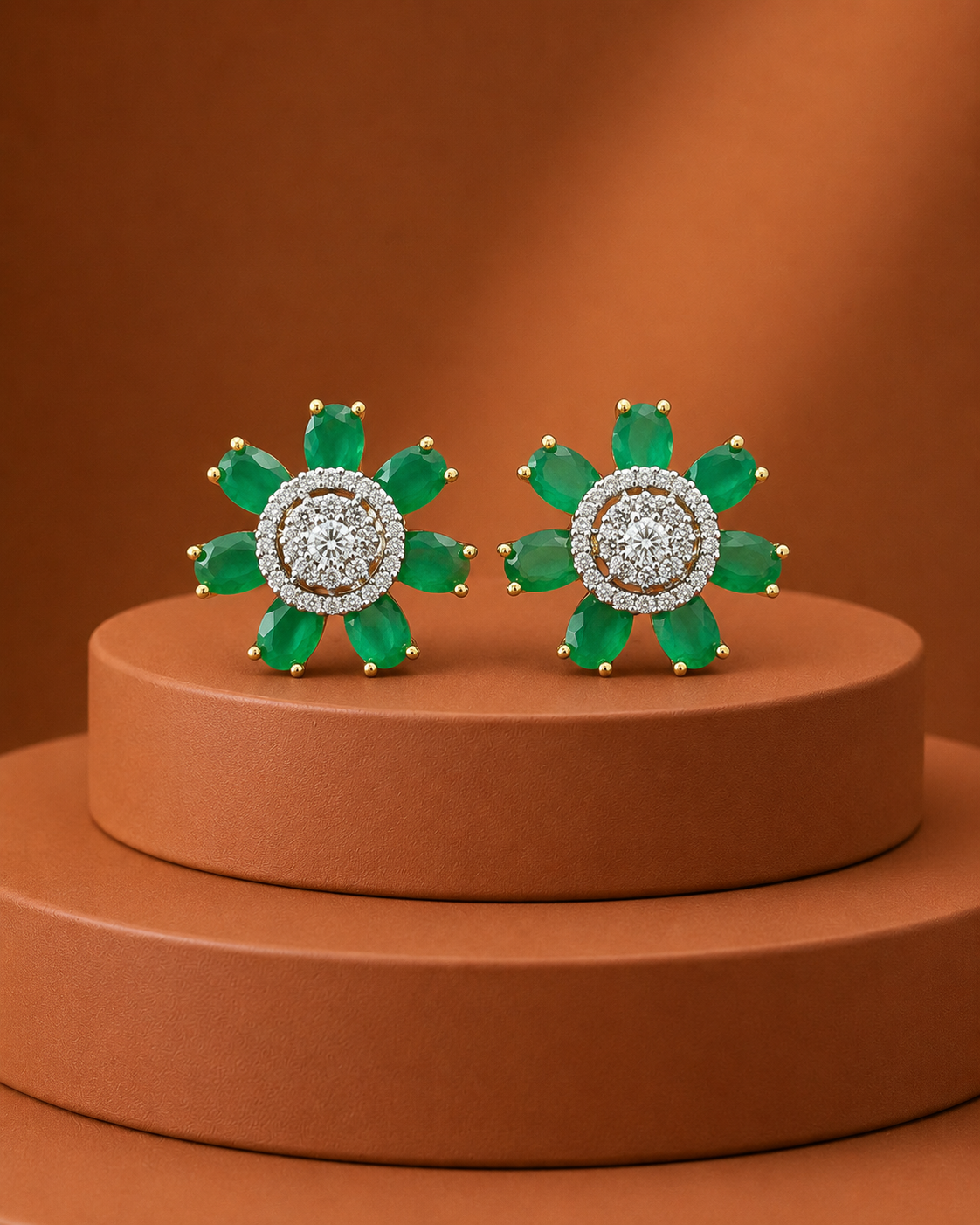 A pair of flower-shaped stud earrings with emerald-cut green stones and a central halo of clear stones, displayed on a round terracotta pedestal against a warm brown background.
