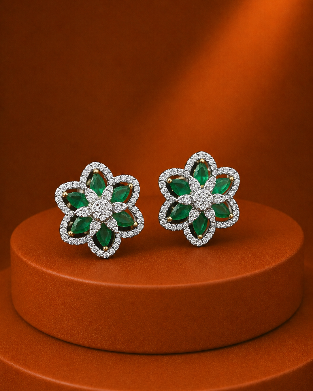 A pair of flower-shaped stud earrings with green emerald-cut stones and silver pavé borders, displayed on a round terracotta pedestal against a warm brown background.