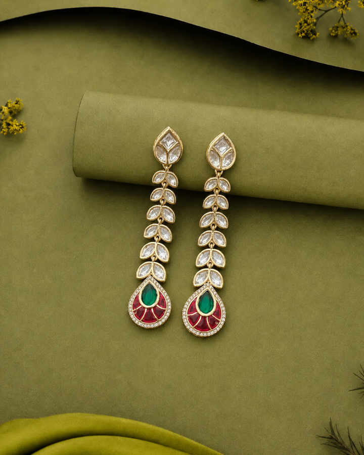 A pair of gold-plated dangle earrings with clear pavé-set stones and green and red enamel details, displayed on an olive green fabric background.