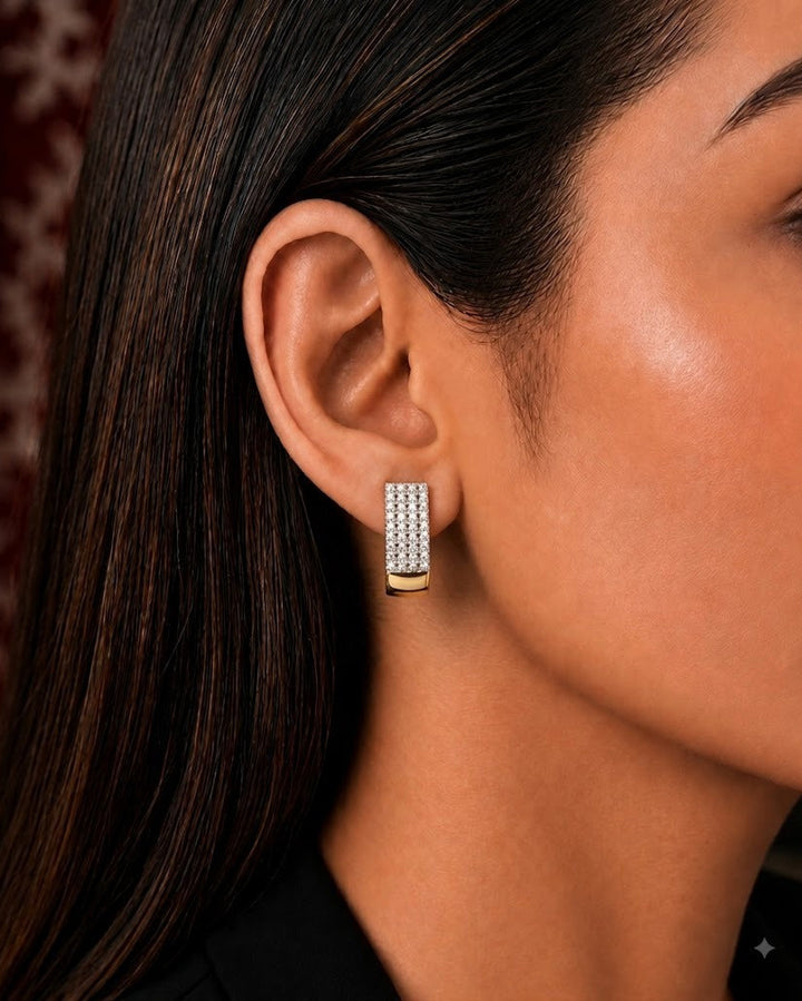 A pair of gold-toned statement earrings displayed on a round orange-brown pedestal.a small rectangular stud earring with pavé-set clear stones and a polished gold band at the bottom.