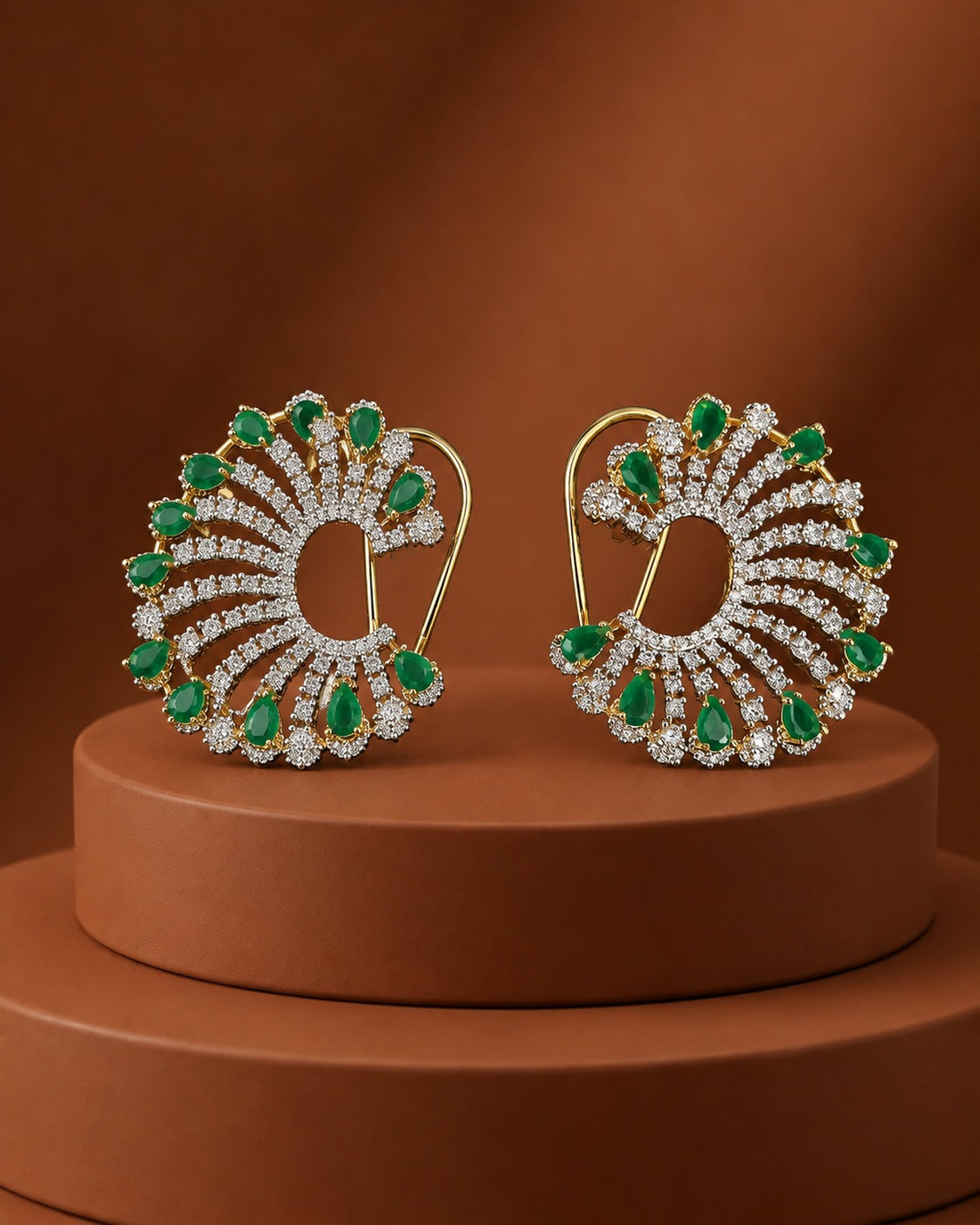A pair of gold-toned drop earrings with fan-shaped clusters of green emerald-cut gemstones and clear stones, displayed on a round brown pedestal against a warm brown background.